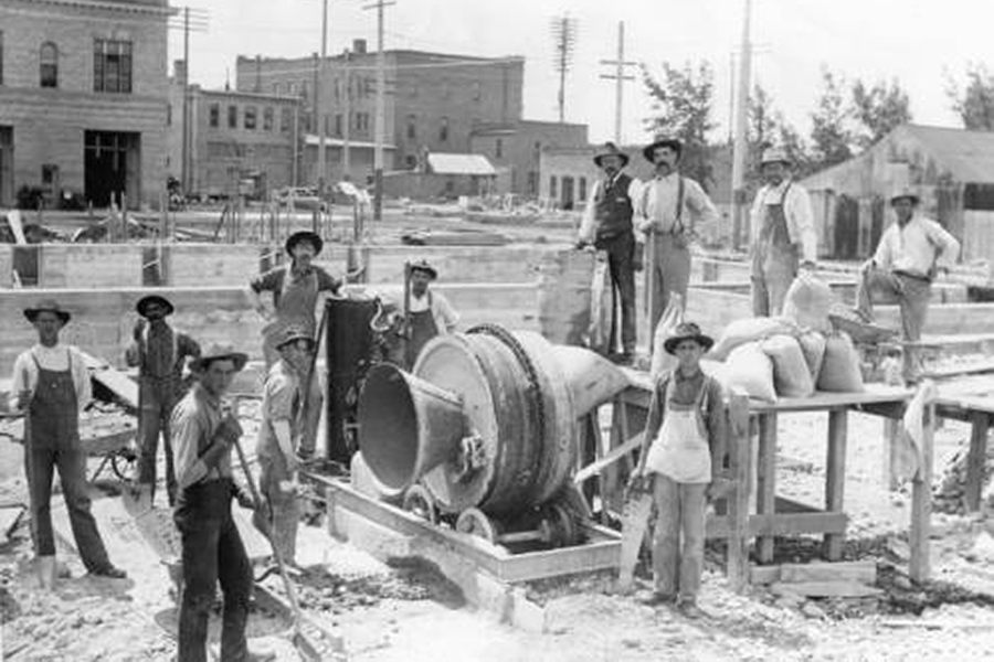 Group of construction workers posing around a vintage cement mixer on an early 20th‑century town construction site.