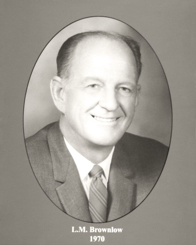 Black-and-white portrait of a smiling man in a suit within an oval frame, captioned'L. M. Brownlow 1970'