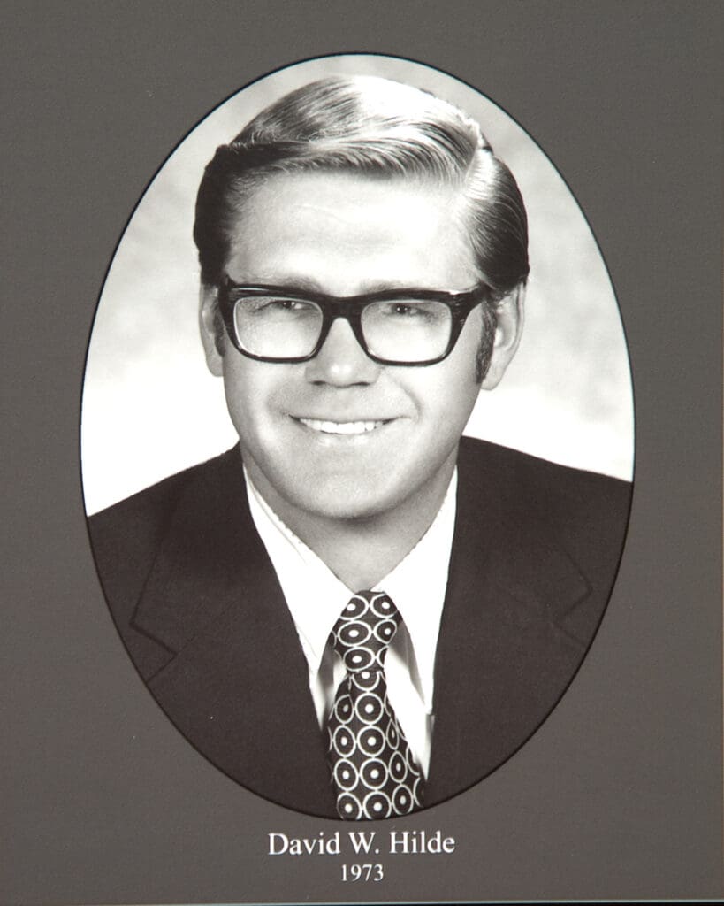 Black-and-white portrait of a man in a suit and patterned tie, framed in an oval on a memorial plaque: David W. Hilde, 1973.