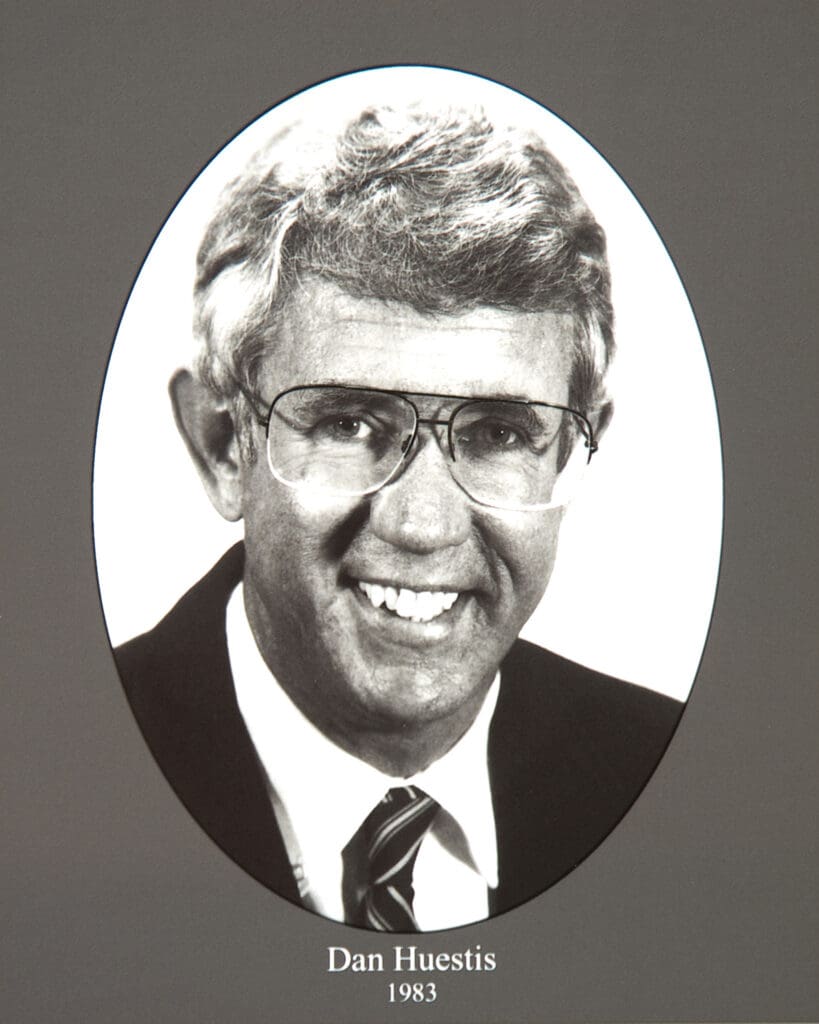 Grayscale portrait of a man in glasses and a suit inside an oval frame, with the caption'Dan Huestis 1983' beneath.