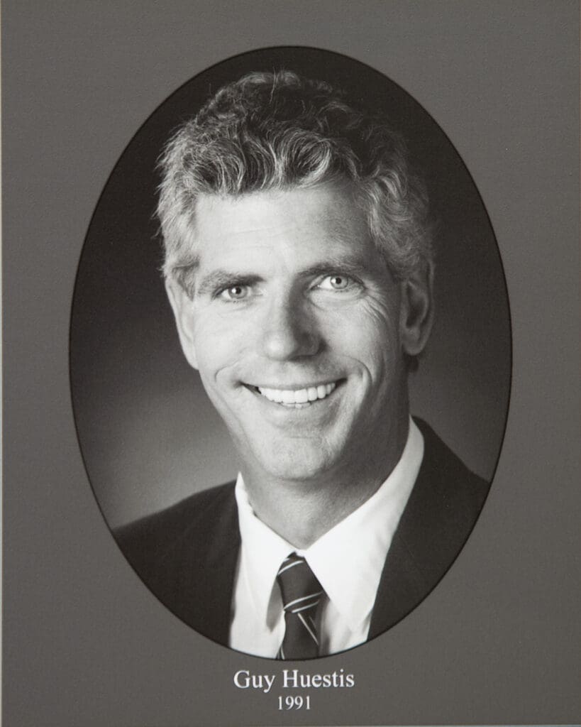 Black-and-white portrait of a smiling man in a suit and tie inside an oval frame, labeled 'Guy Huestis 1991'