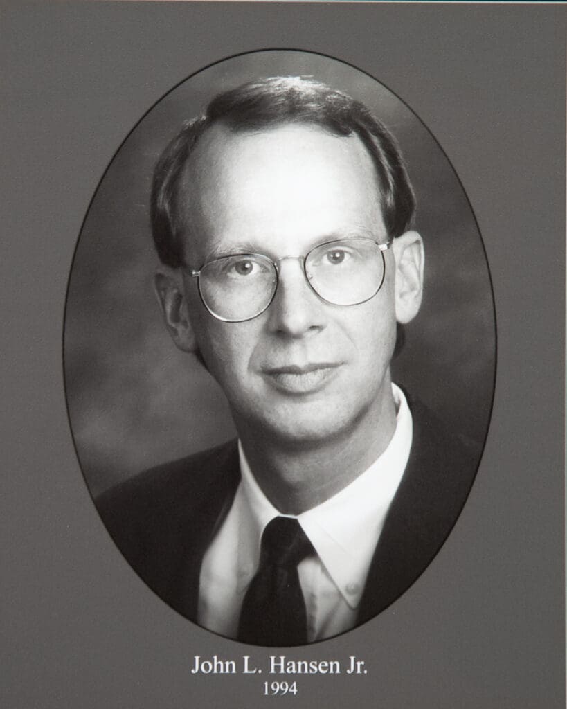 Black-and-white oval-framed portrait of John L. Hansen Jr. wearing glasses, a suit, and tie; caption reads'John L. Hansen Jr. 1904–1994'.