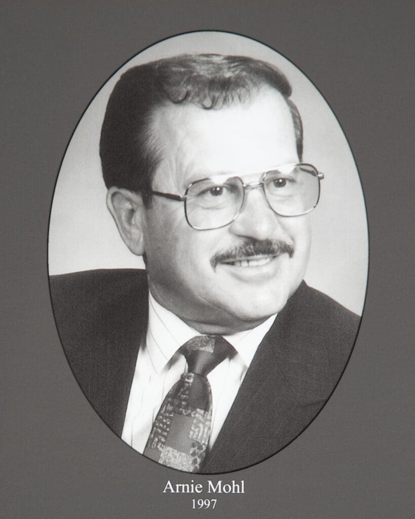Oval-framed black-and-white portrait of a man wearing glasses and a suit, labeled Arnie Mohl, 1997.