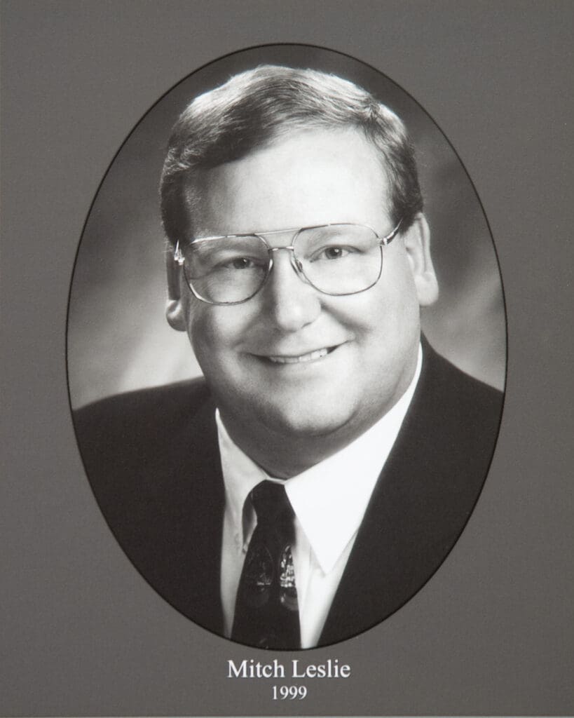 Black-and-white portrait of Mitch Leslie wearing glasses and a suit, framed in an oval; caption reads 'Mitch Leslie 1999'