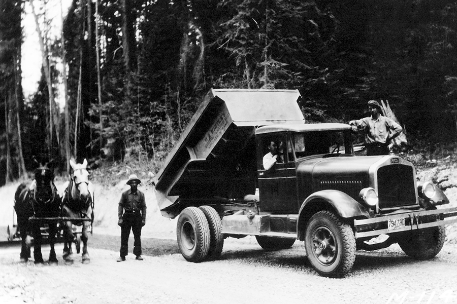 Black-and-white photo of a vintage dump truck unloading on a dirt road, with a man beside and a man in the cab, while a horse-drawn wagon and team stand in the background.
