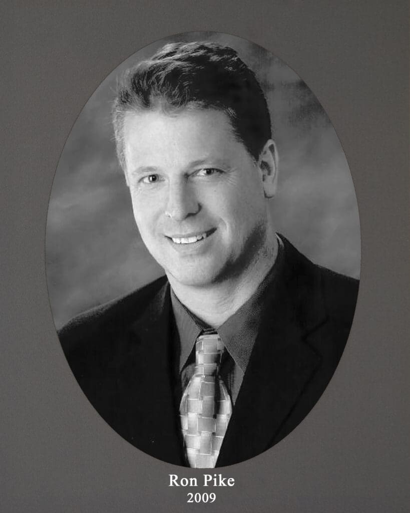 Portrait of a smiling man in a suit inside an oval frame on a gray background; caption reads'Ron Pike' with year '2009' beneath.