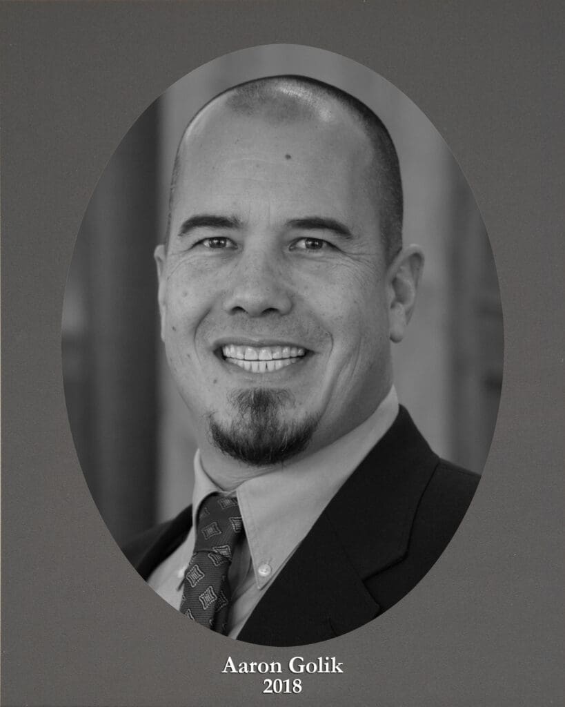 Black-and-white portrait of Aaron Golik in a suit and tie, smiling; caption reads'Aaron Golik 2018' at the bottom.