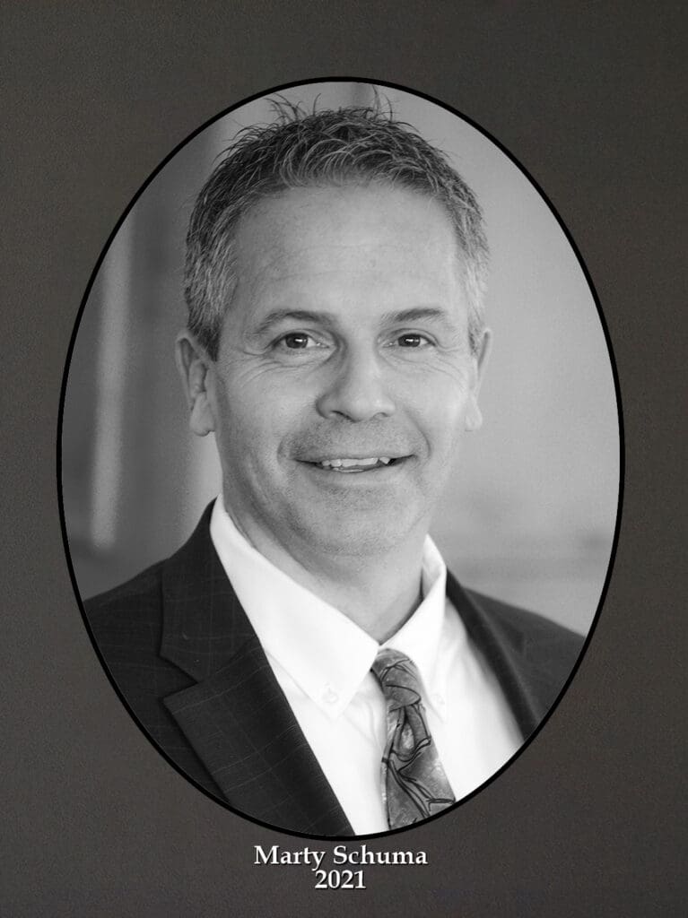 Oval-framed black-and-white portrait of a smiling man in a suit and tie, with caption 'Marty Schuma 2021'.