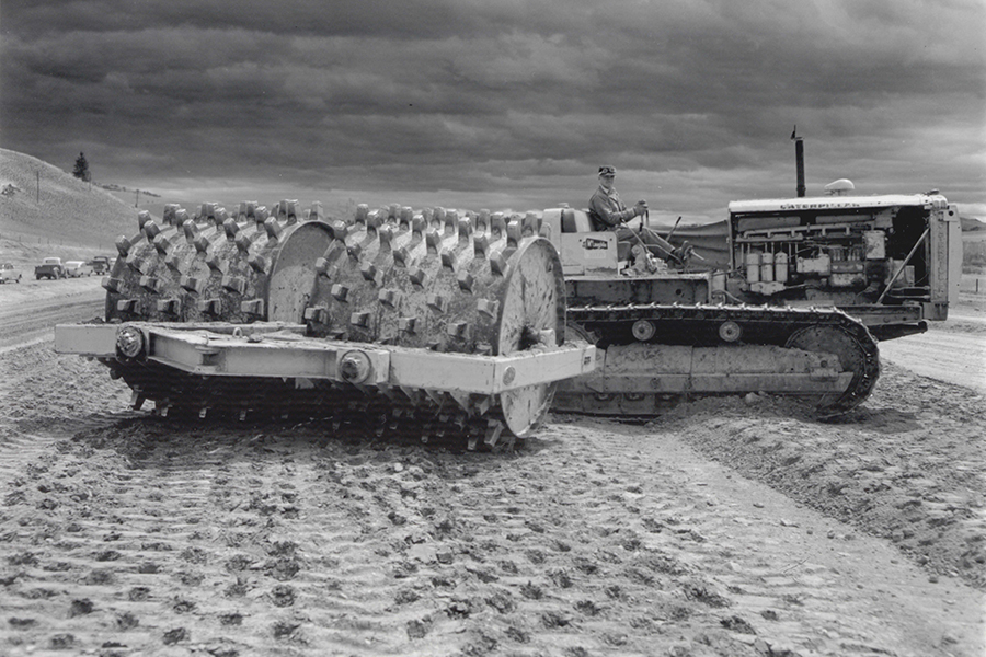 Black-and-white photo of a large tractor with a spiked roller attachment used for soil compaction on a construction site, under a cloudy sky, with a worker seated at the controls. - concise informative alt text 1