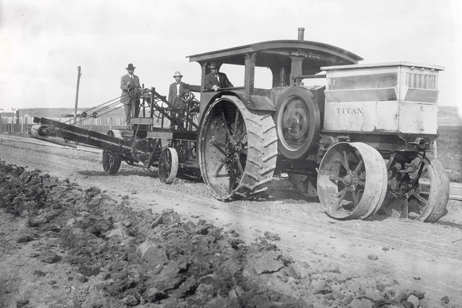 Vintage Titan tractor with large metal wheels pulling a plow on a dirt rural road, four men in hats standing nearby.