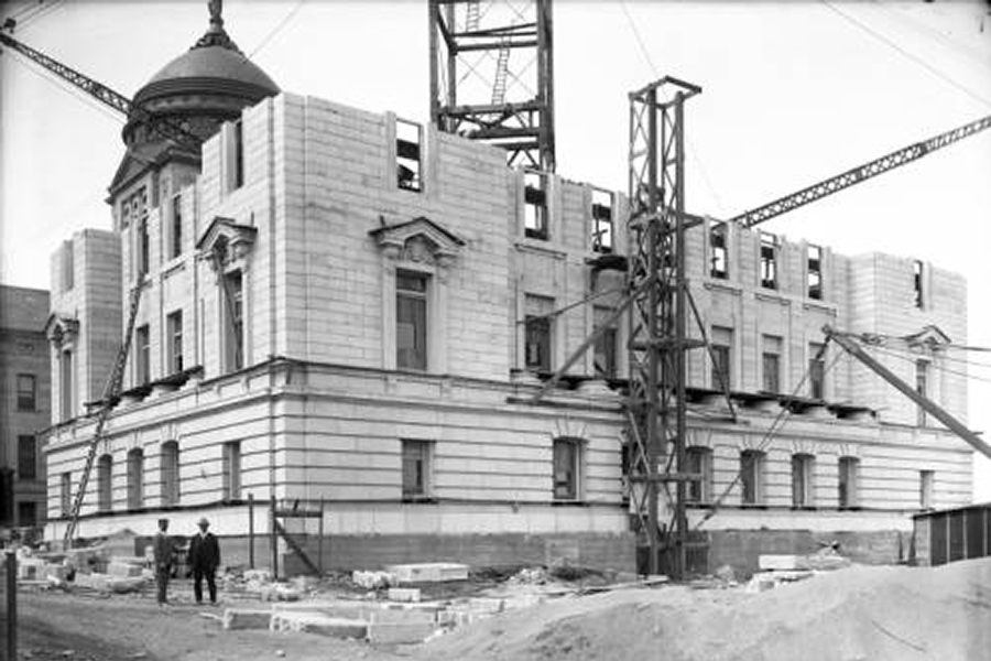 Stone building under construction with a domed turret, scaffolding, and tall cranes; two workers stand in the foreground.