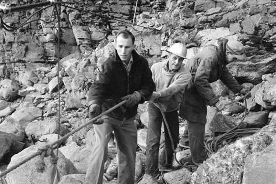 Three men work in a rocky excavation, using shovels and a rope in a quarry setting (black and white).