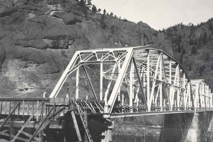 Black-and-white photo of a steel truss bridge spanning a river with rocky hills in the background, likely historic infrastructure.
