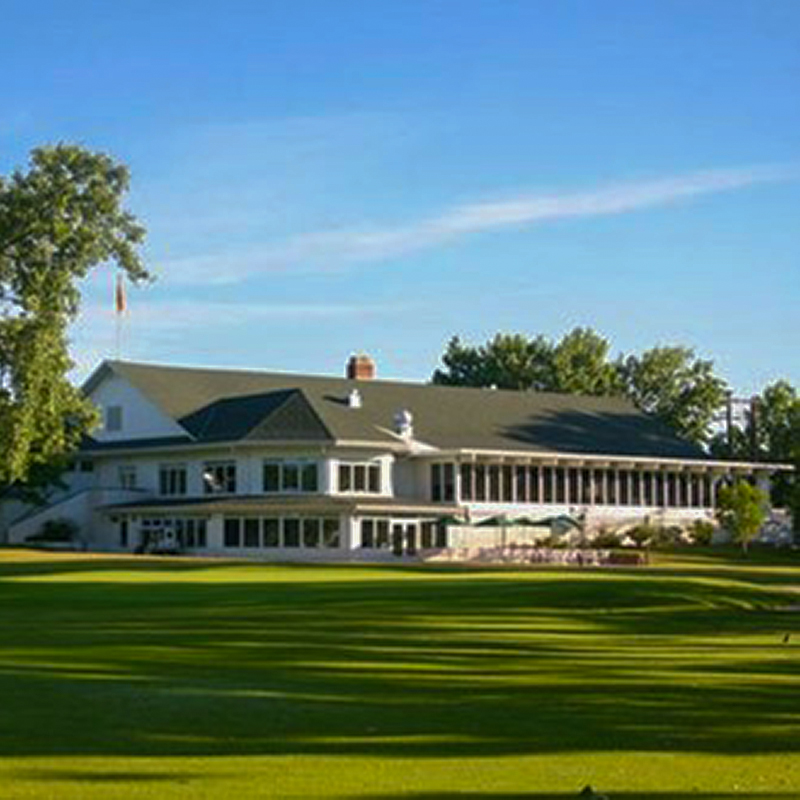White clubhouse with a dark roof on a sunny golf course, surrounded by trees and a green lawn.