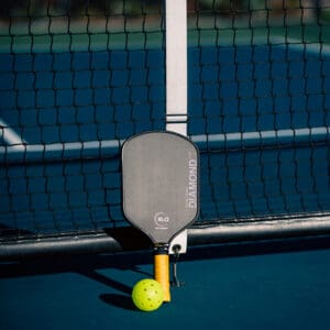 Padel racket leaning against a tennis net with a neon yellow-green ball on a blue court.