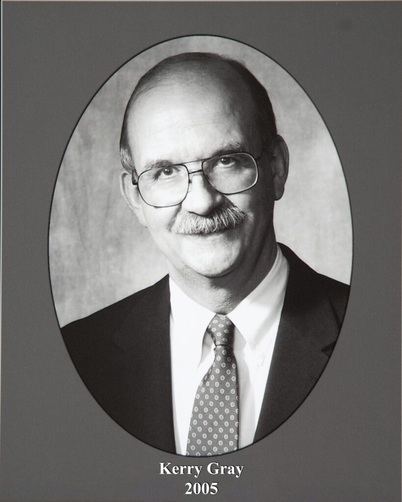 Grayscale portrait of a man in a suit and tie inside an oval frame, with the caption 'Kerry Gray 2005' beneath.