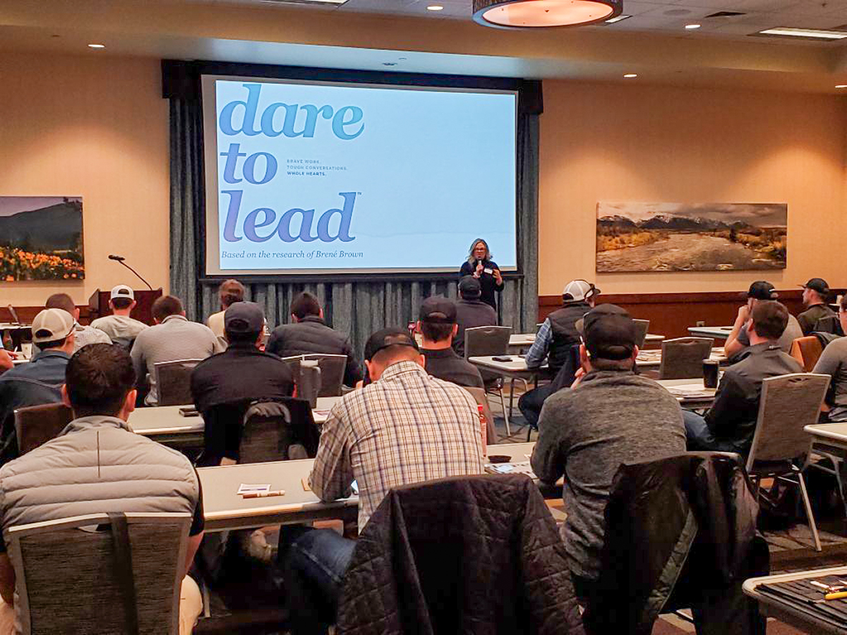 Speaker presenting on a stage in a conference room with a large screen that reads 'dare to lead' and an attentive audience seated at tables behind.