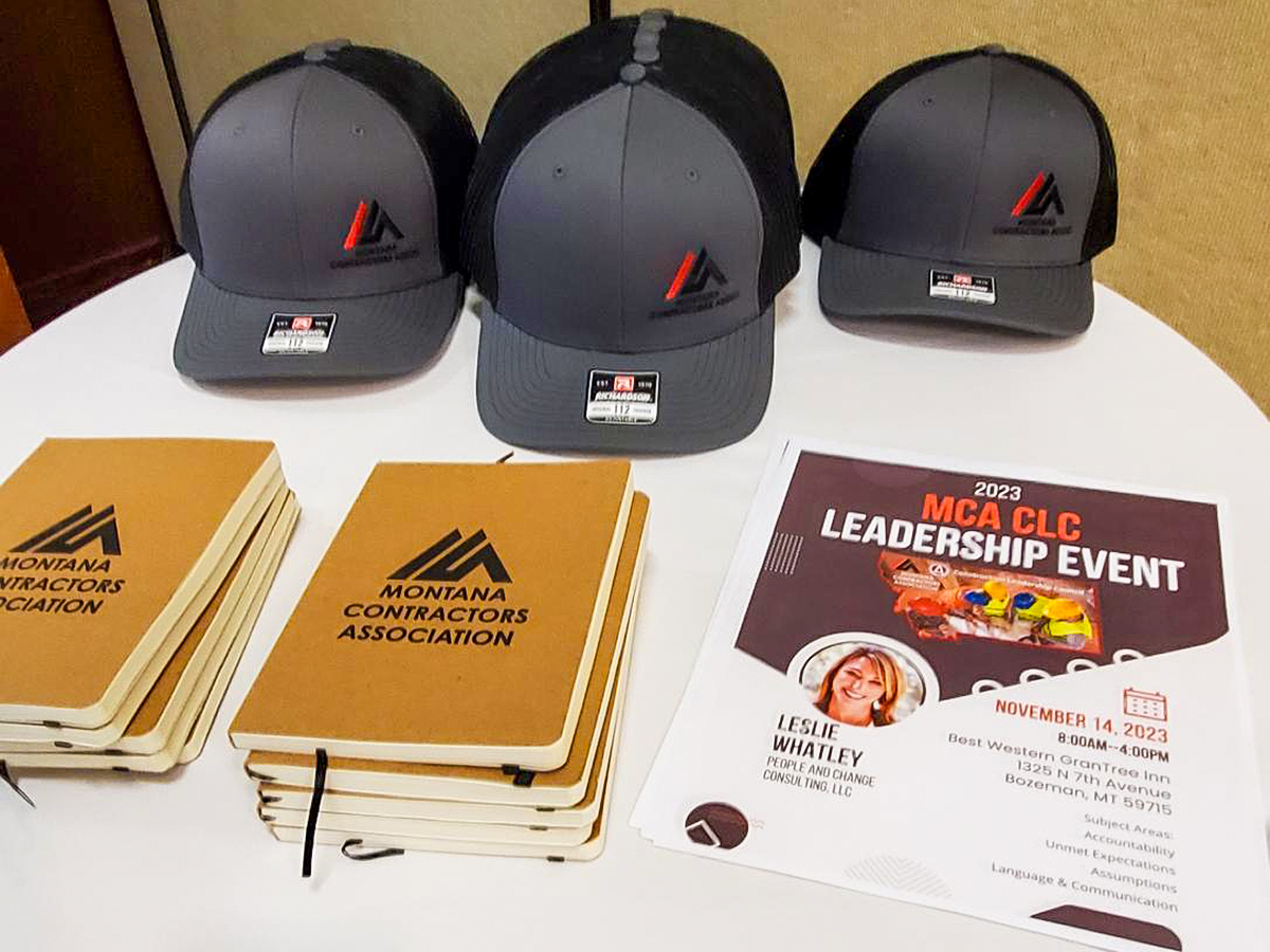Hats and notebooks labeled Montana Contractors Association arranged on a table beside a Leadership Event flyer (2023 MCA CLC).